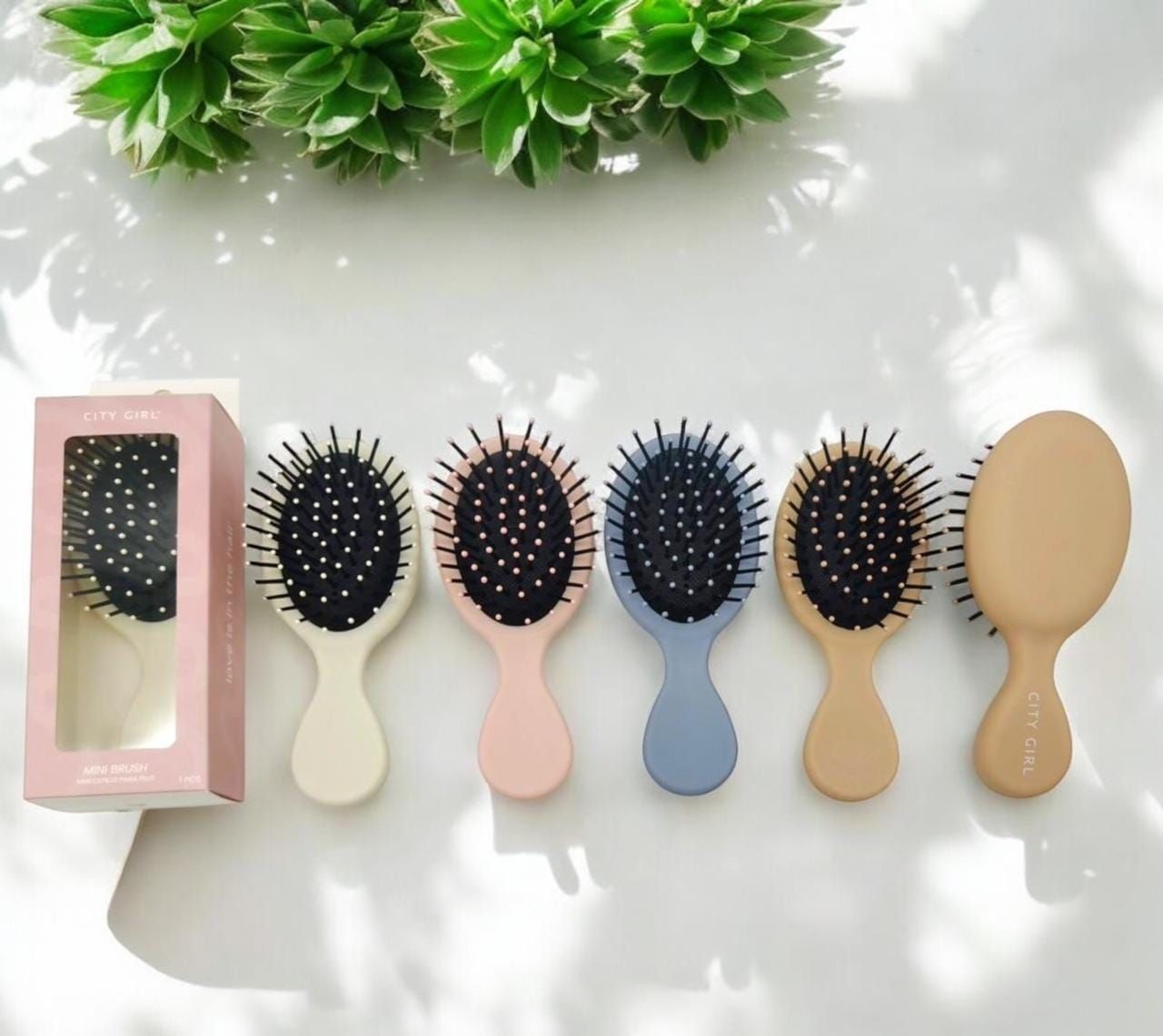 Cinco mini cepillos para el cabello de colores pastel en fila, uno en una caja, fondo de planta verde.
