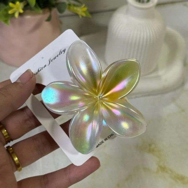 Mano sosteniendo una pinza para el cabello con una flor iridiscente en una etiqueta, con un anillo dorado y flores de fondo.