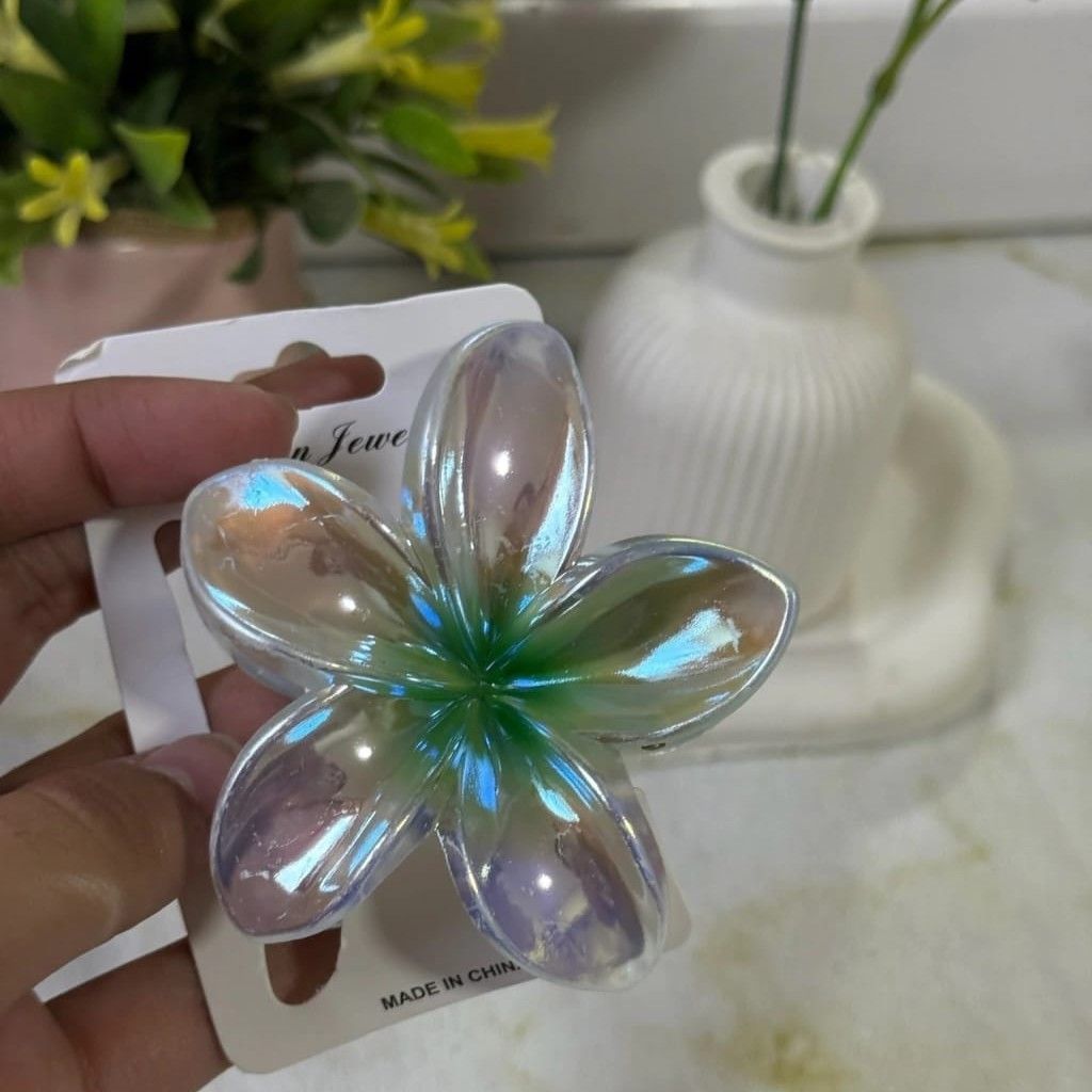 Una mano sostiene una horquilla iridiscente con forma de flor sobre una tarjeta. Al fondo, un jarrón con flores.