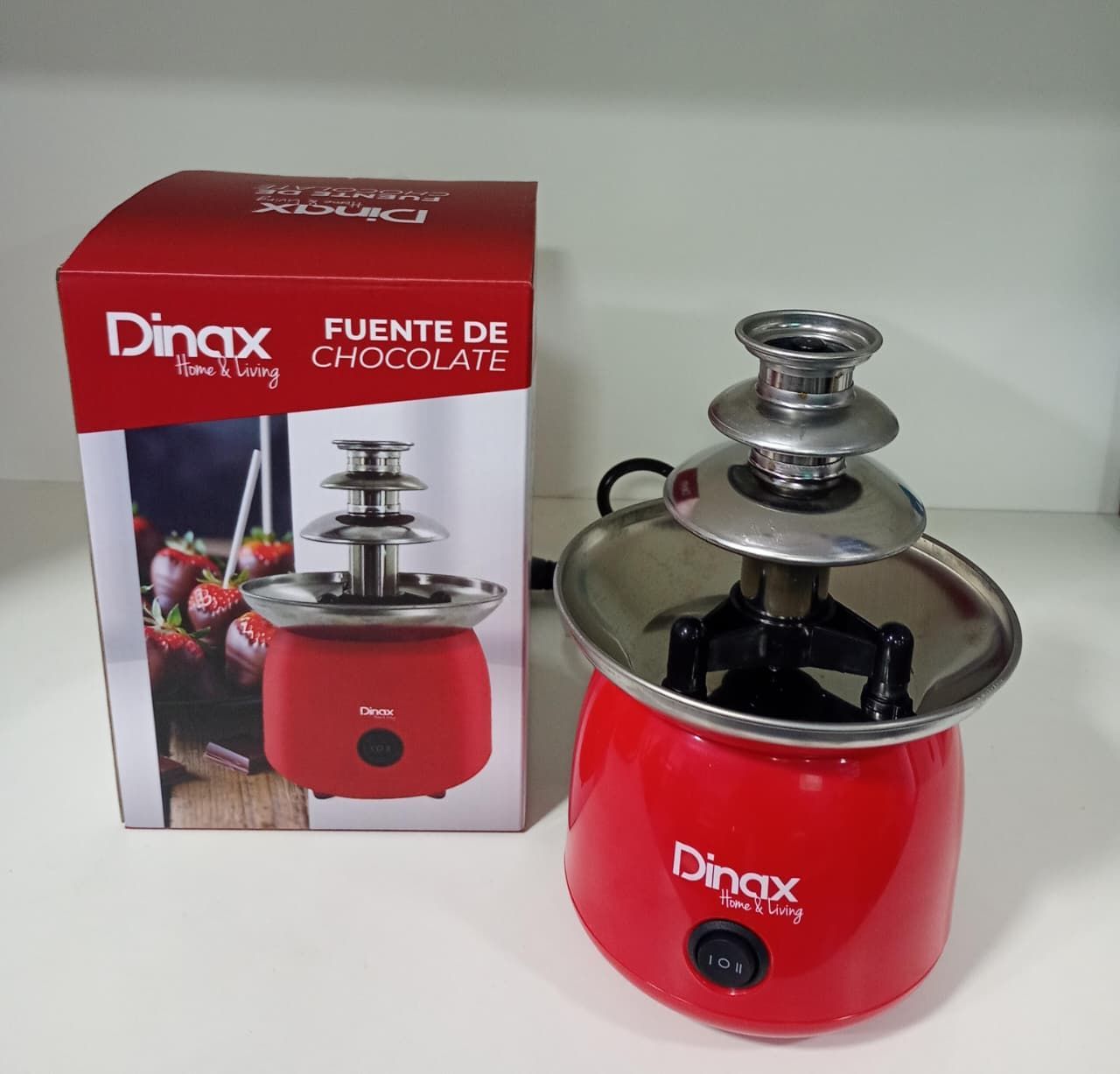 Fuente de chocolate Dinax roja junto a su caja, con fresas cubiertas de chocolate sobre la caja.