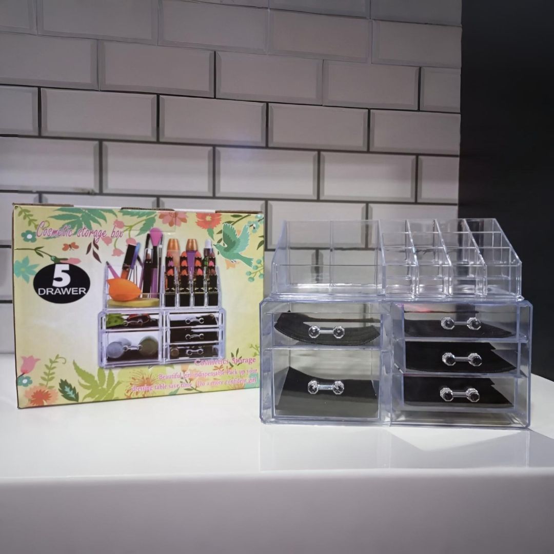 Un organizador de maquillaje de acrílico transparente con cajones, al lado de su caja, contra una pared de azulejos blancos.