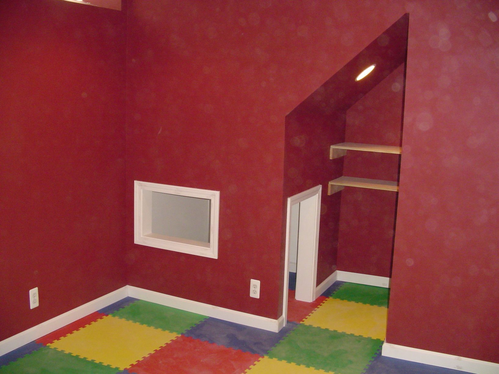 Red-walled playroom with colorful foam tiles, a small window, and a built-in play area with shelves.
