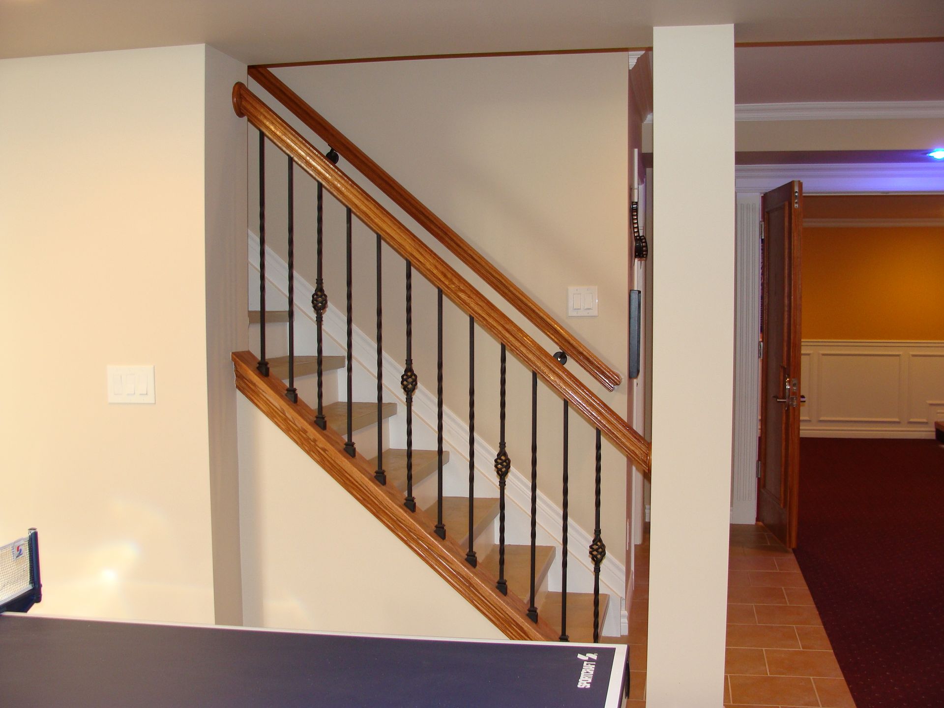Staircase with wooden handrails, black iron spindles, beige walls, and doorway to a room with red carpet.