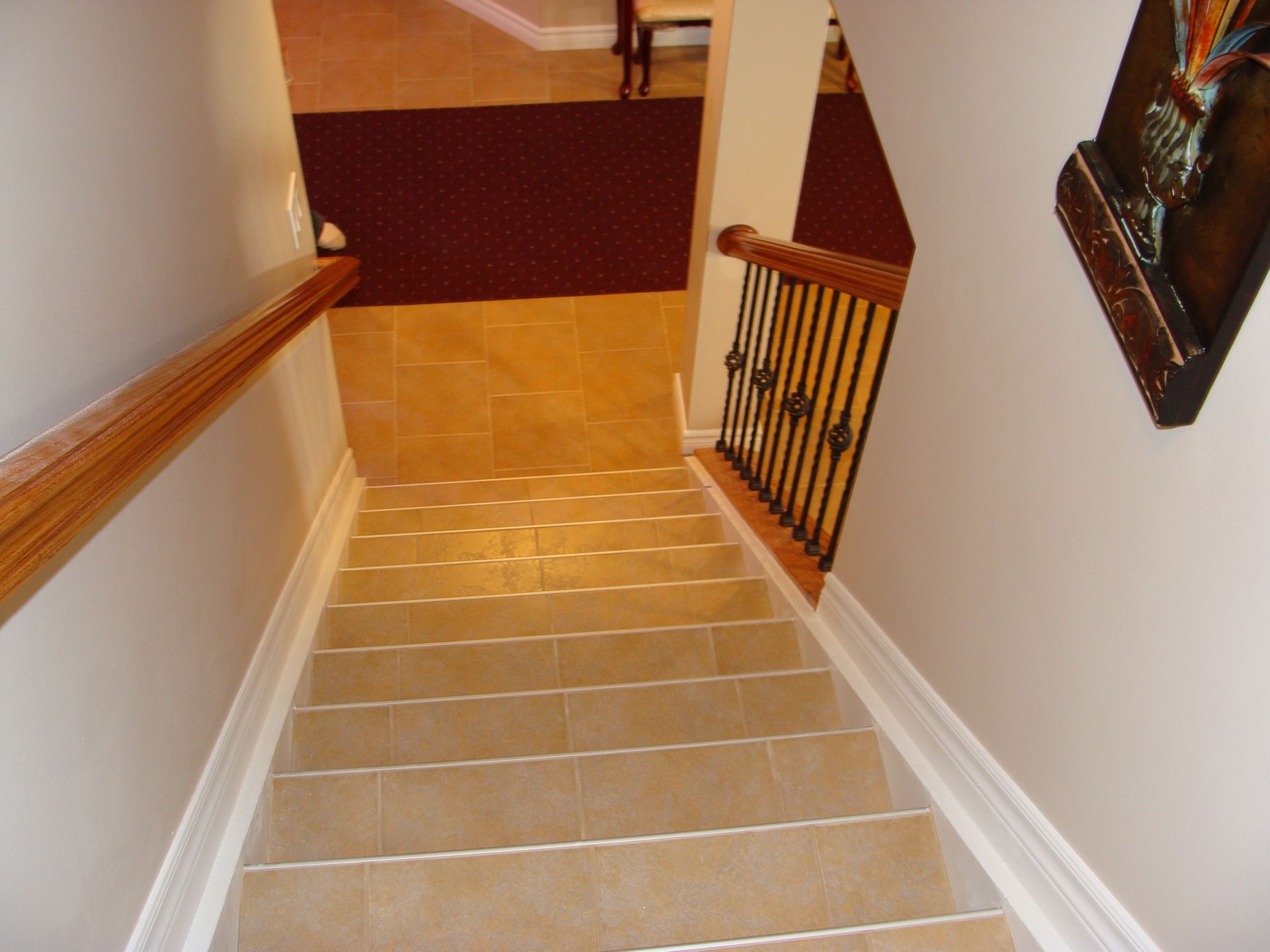 Staircase descending from a hallway. Beige tiled steps, brown carpet, wood railing, white walls.