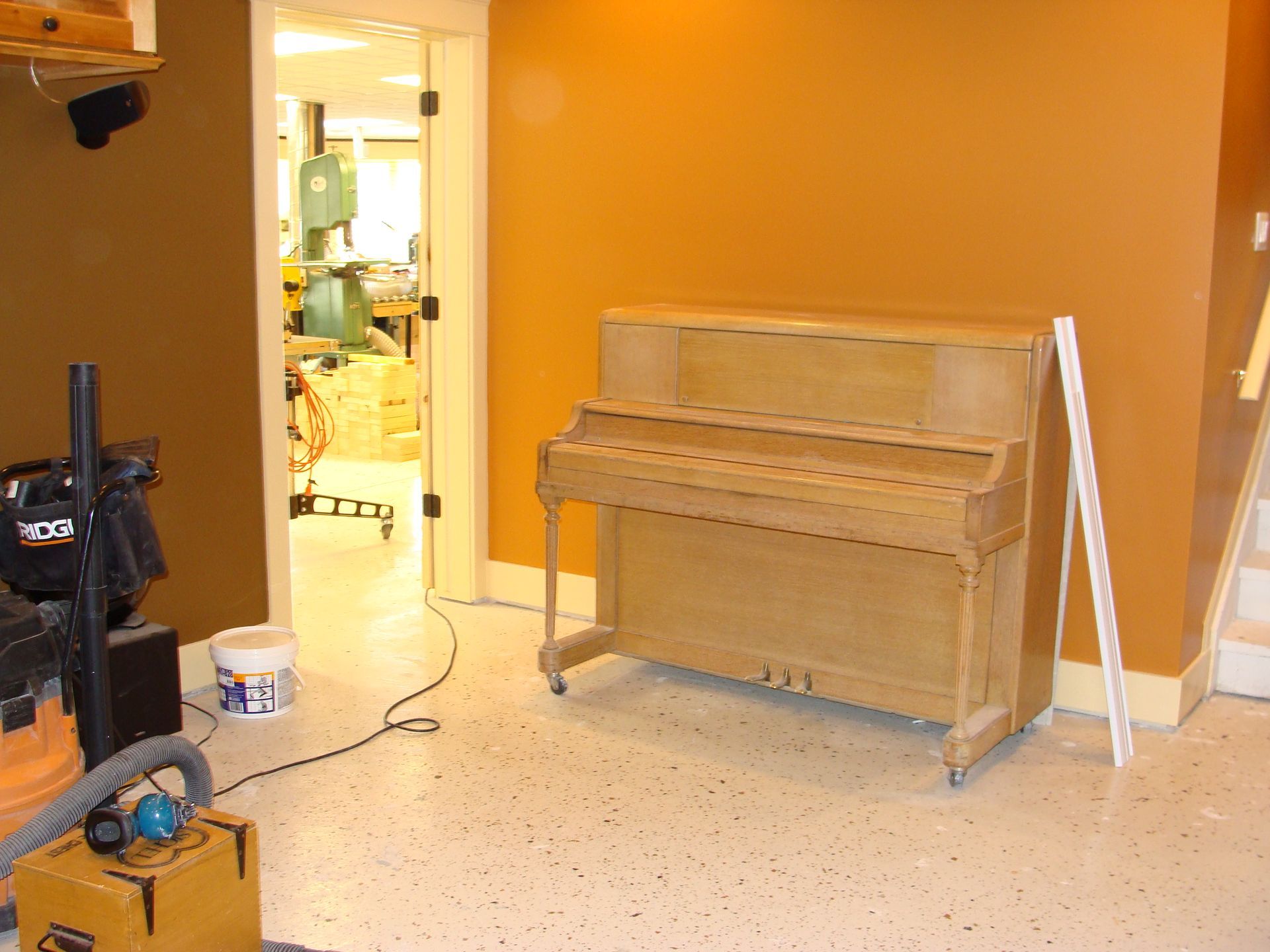A piano against an orange wall in a room with tools and a doorway.