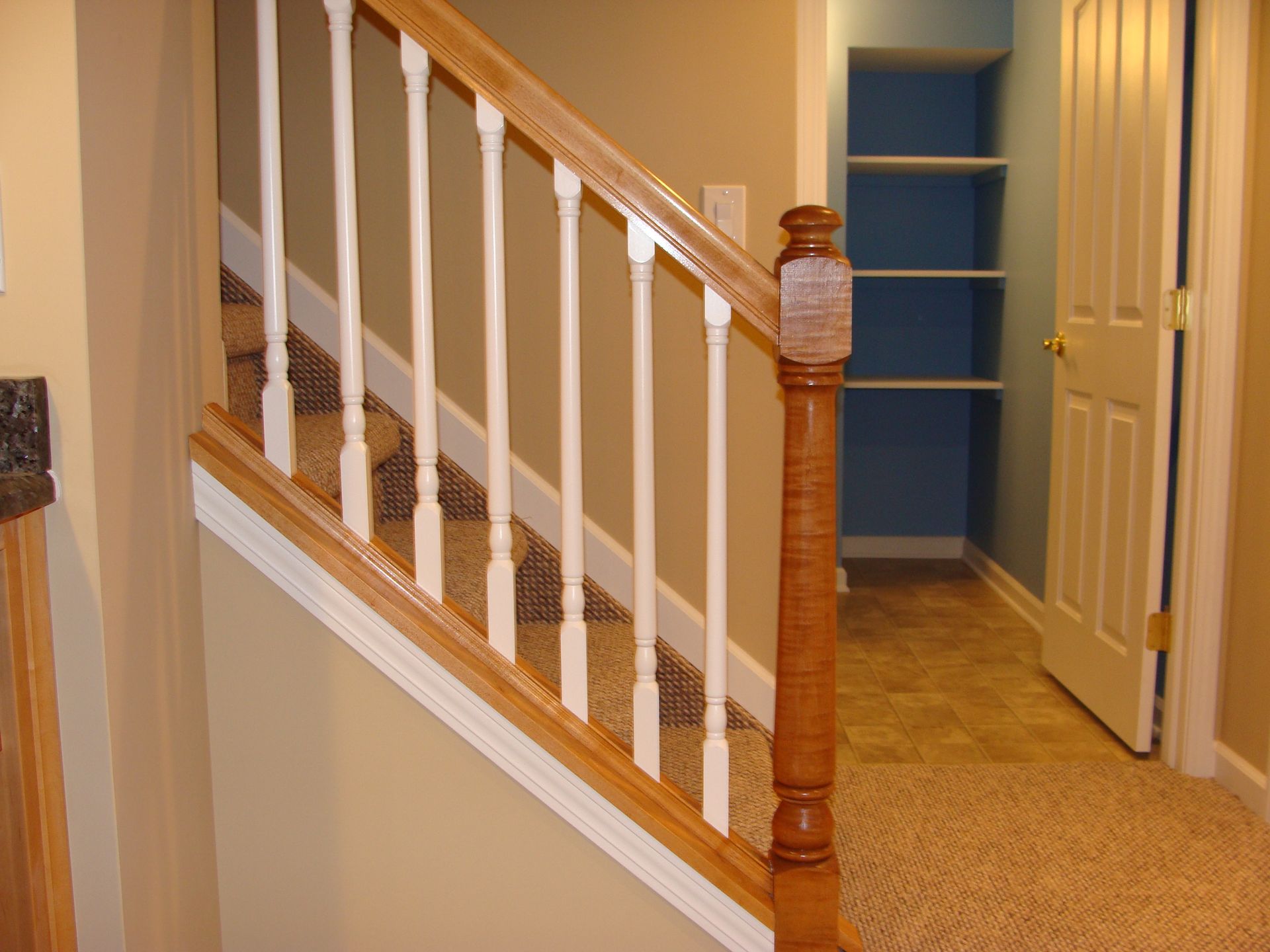 Staircase with wood handrail and white spindles, leading to a hallway with shelves and a door.