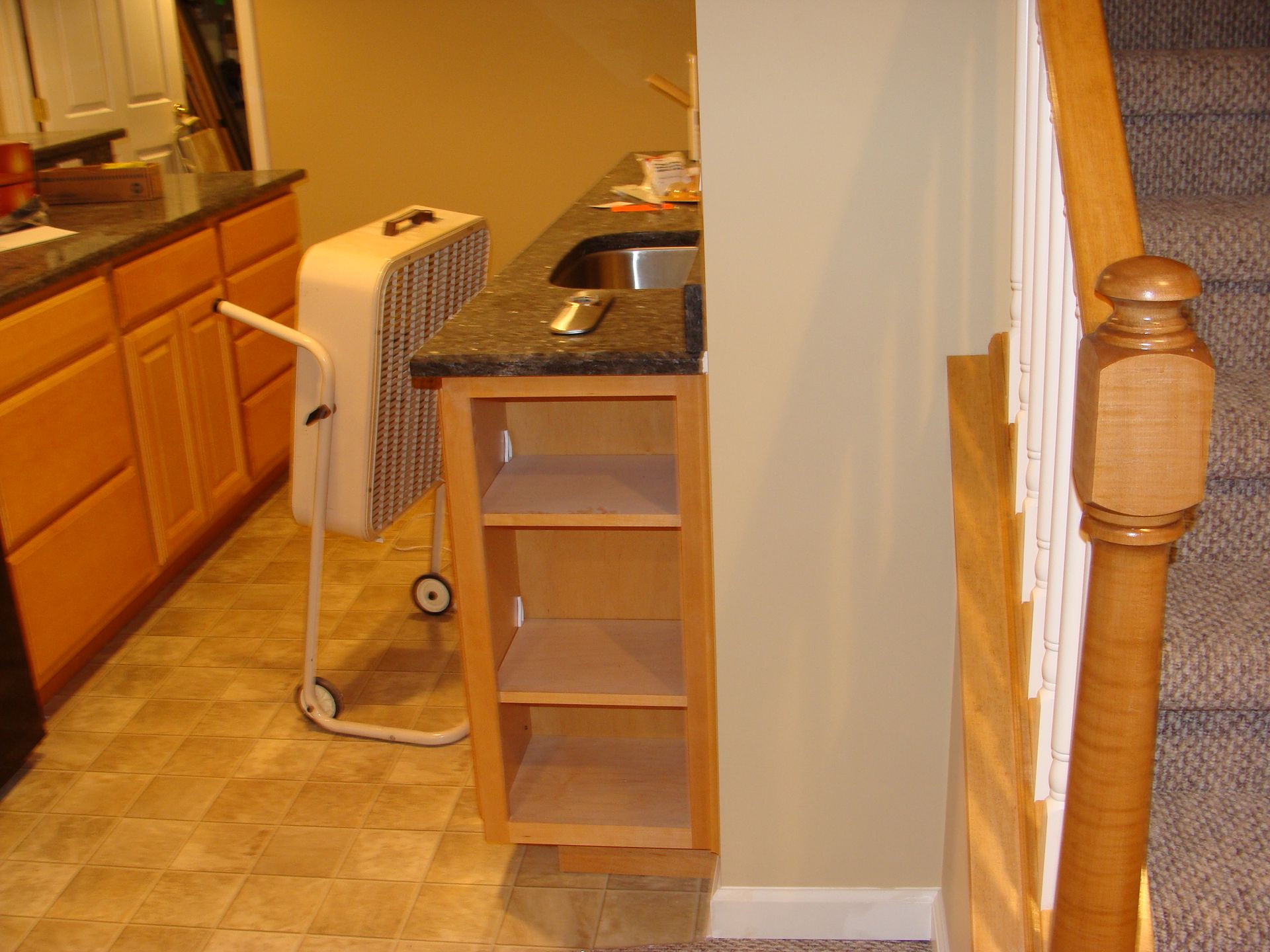Kitchen counter with sink and open shelving next to a staircase; a fan is nearby.