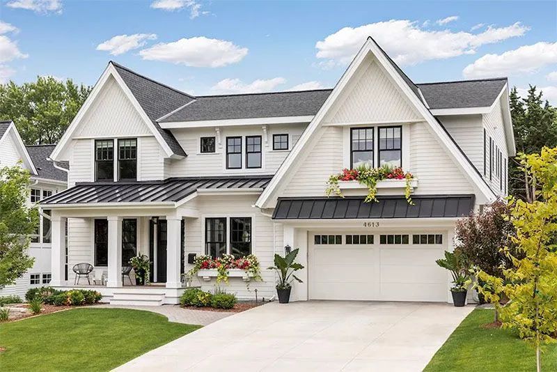 White two-story house with black roof, garage, and windows. Green lawn, flower boxes, and blue sky.