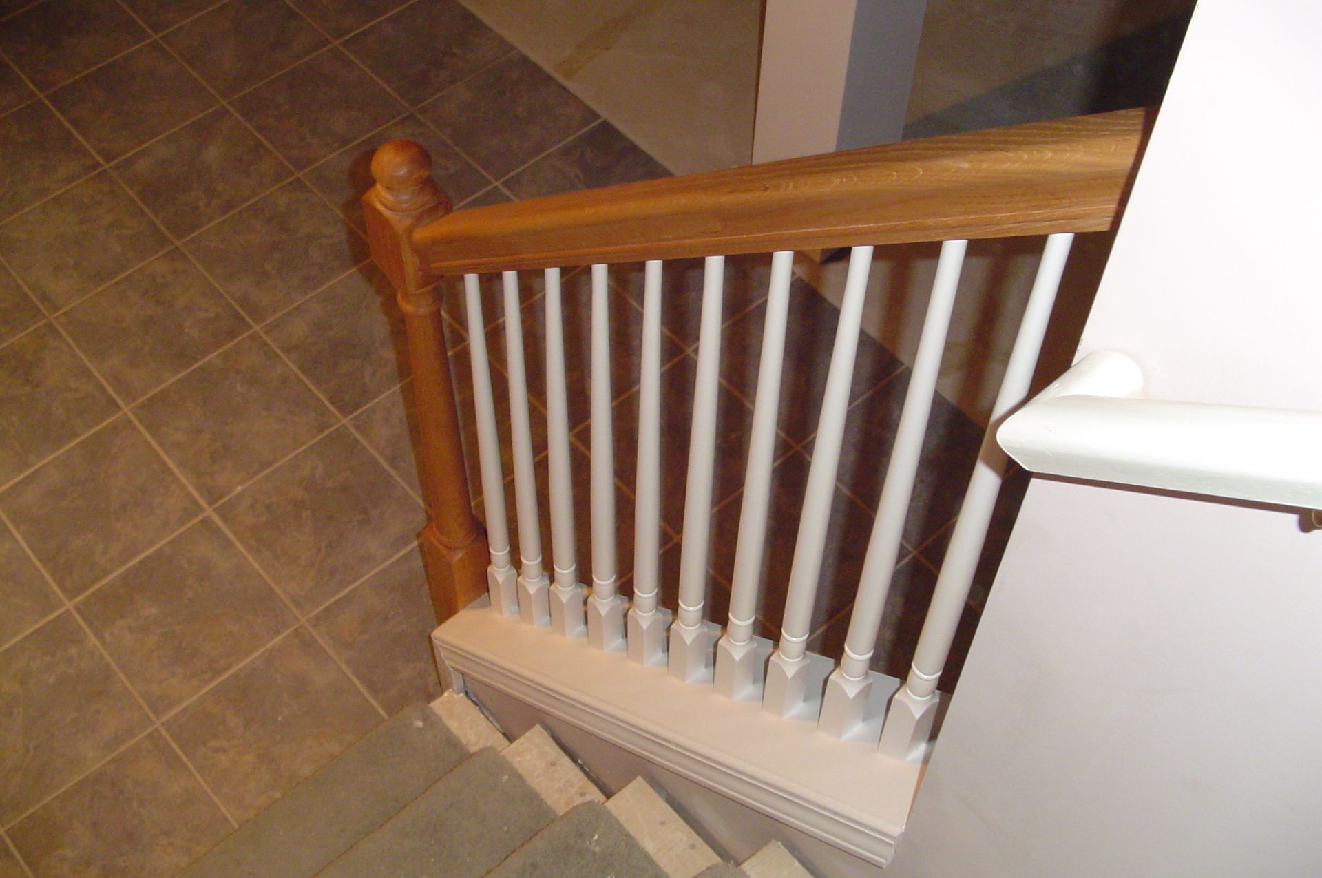 Staircase with wooden handrail and white spindles, leading down into a room with tile flooring.