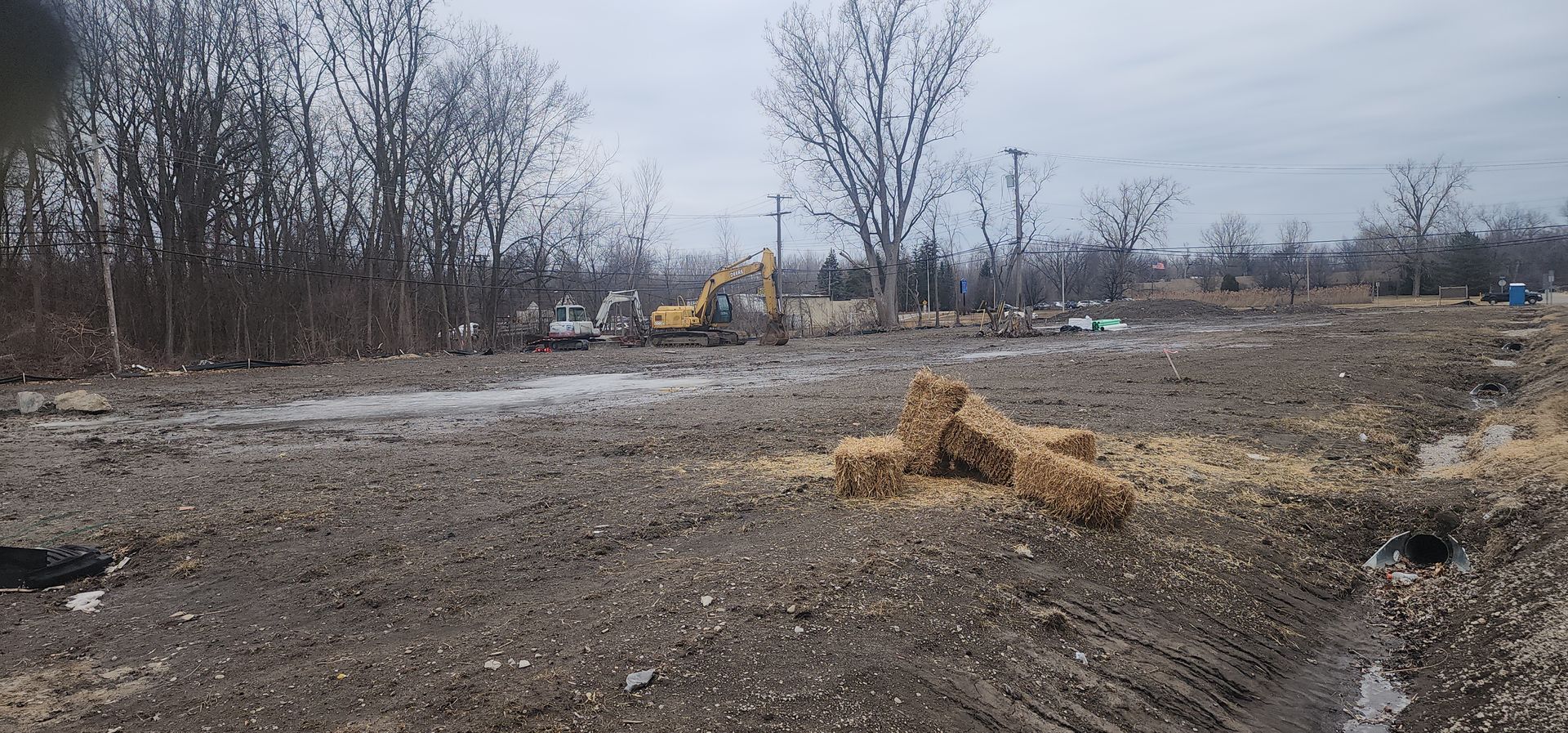 Construction site with mud, trees, and an excavator, some hay bales in the foreground.