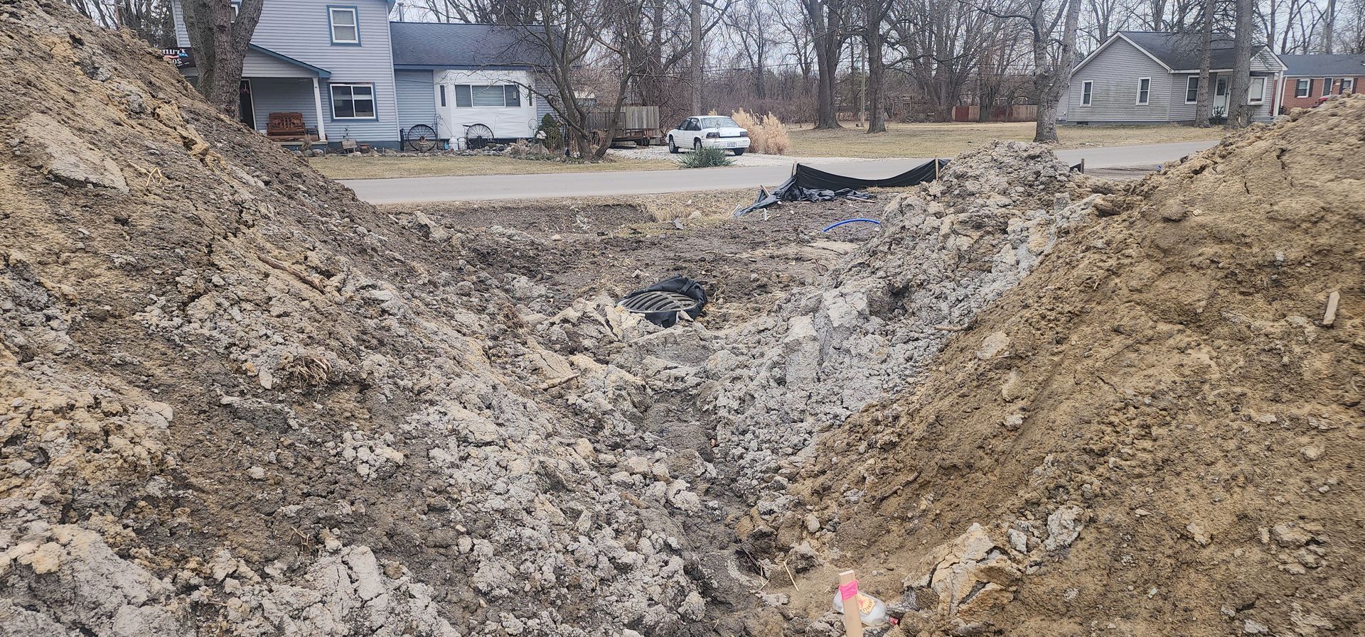 Construction site with large piles of dirt and a ditch in front of a house and road.