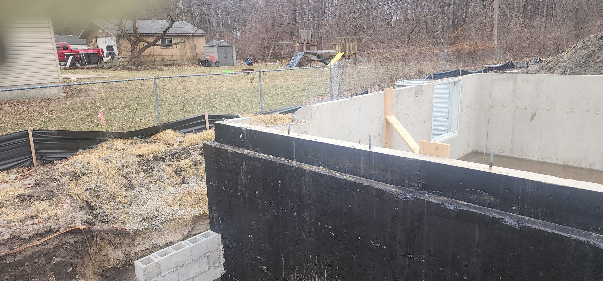 Construction site with concrete foundation, black wall, and grassy area in the background.