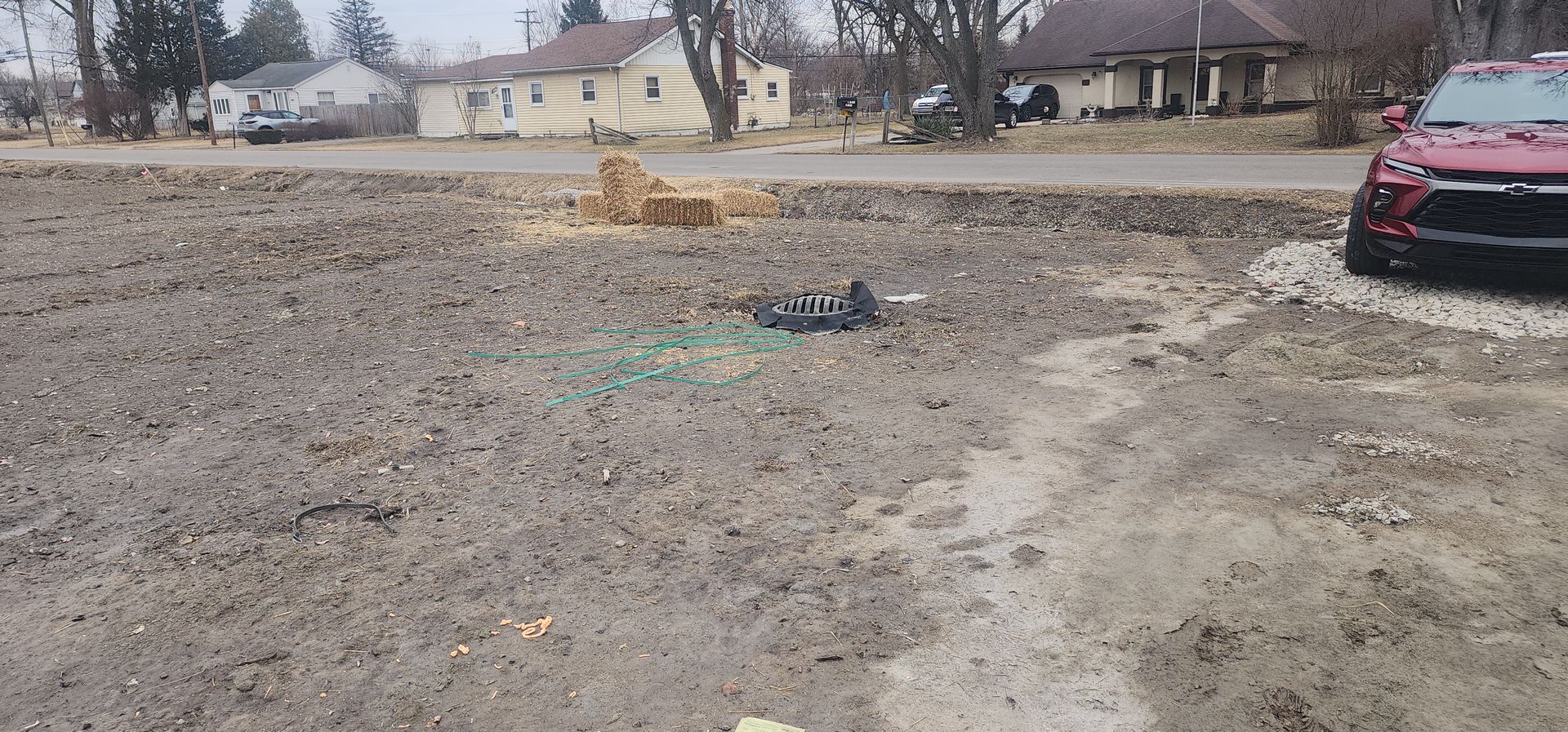 A mostly bare lot with a red car parked at the right. Houses line the road in the background.