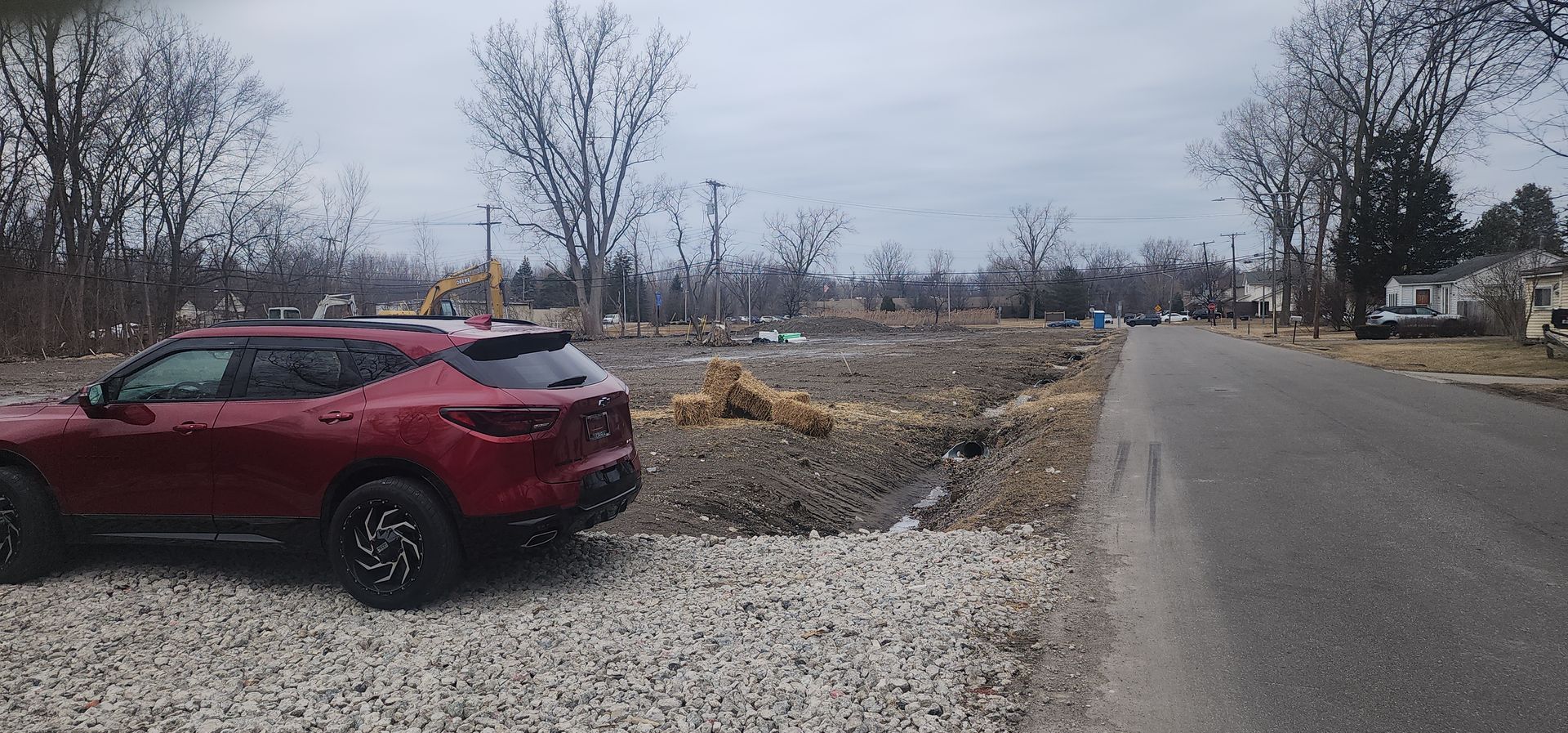 Red SUV parked on a gravel lot next to a road. Brown dirt and bare trees in the background. Cloudy sky.