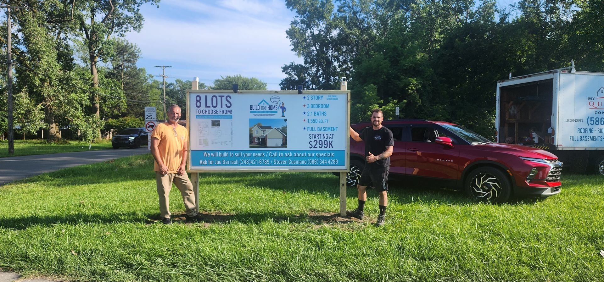 Two men stand by a sign in front of a red car and a truck. Sign is for 8 lots.
