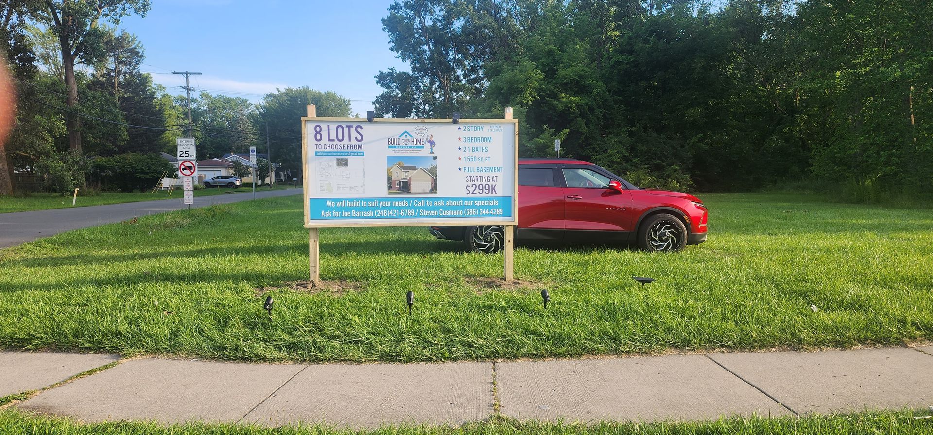 A red car parked next to a sign in a grassy area with a sidewalk.