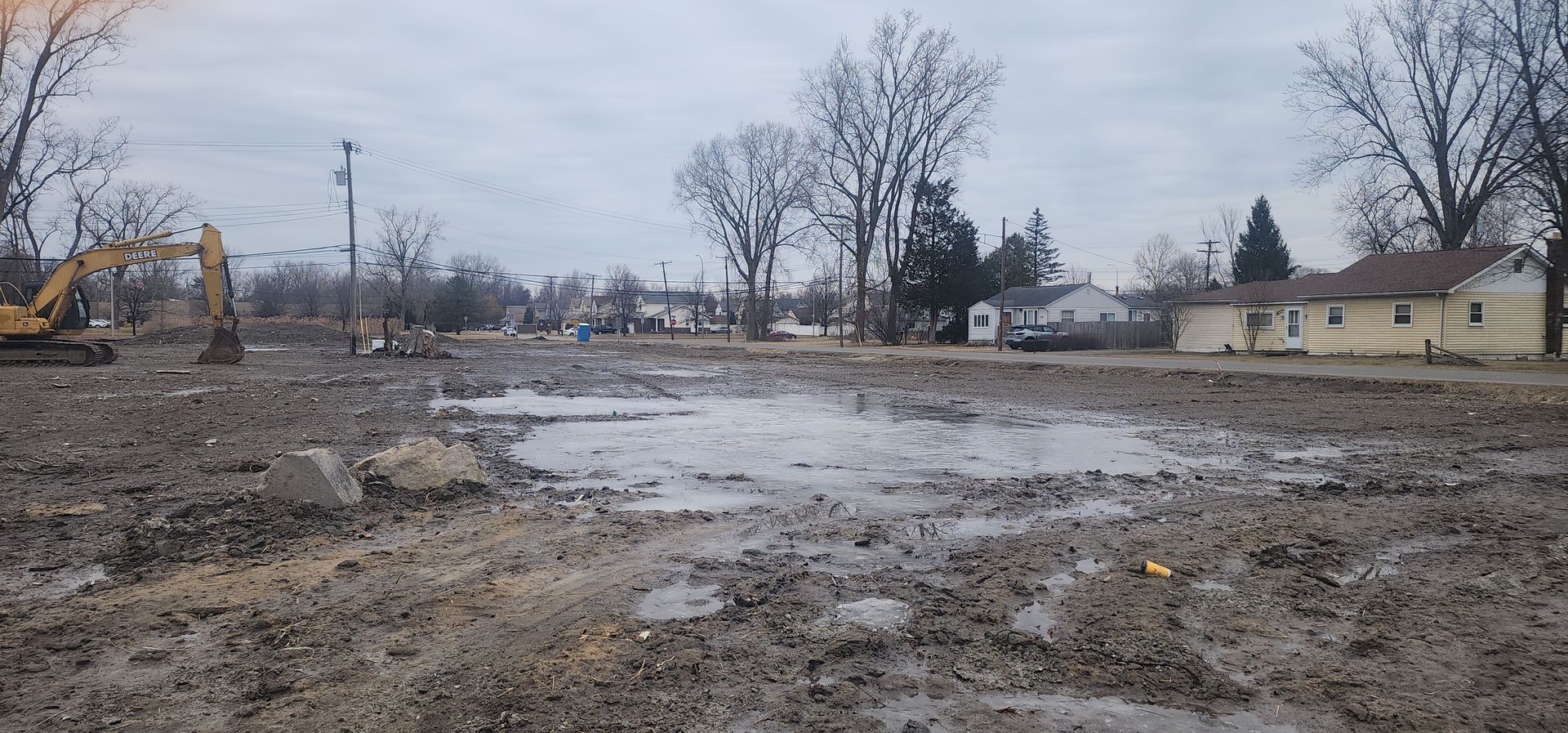 A muddy construction site with an excavator, puddles, and houses in the distance under a cloudy sky.