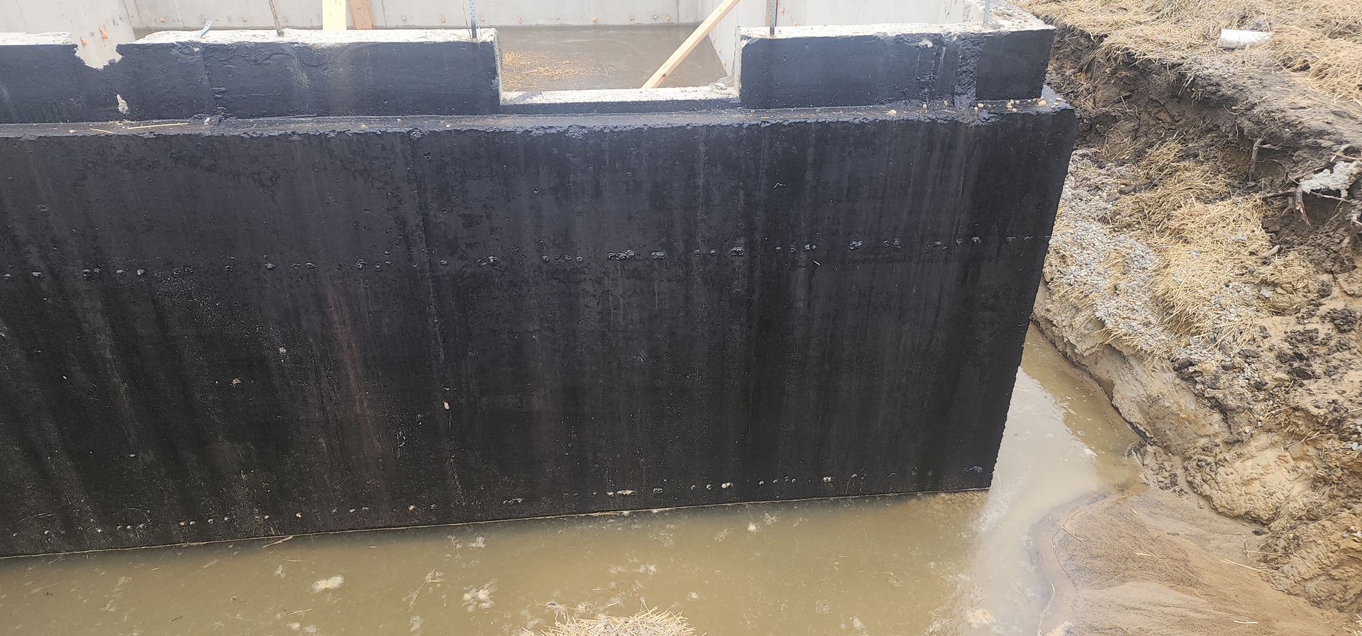 Black coated concrete wall, likely a foundation, in muddy water. The ground around is also muddy.