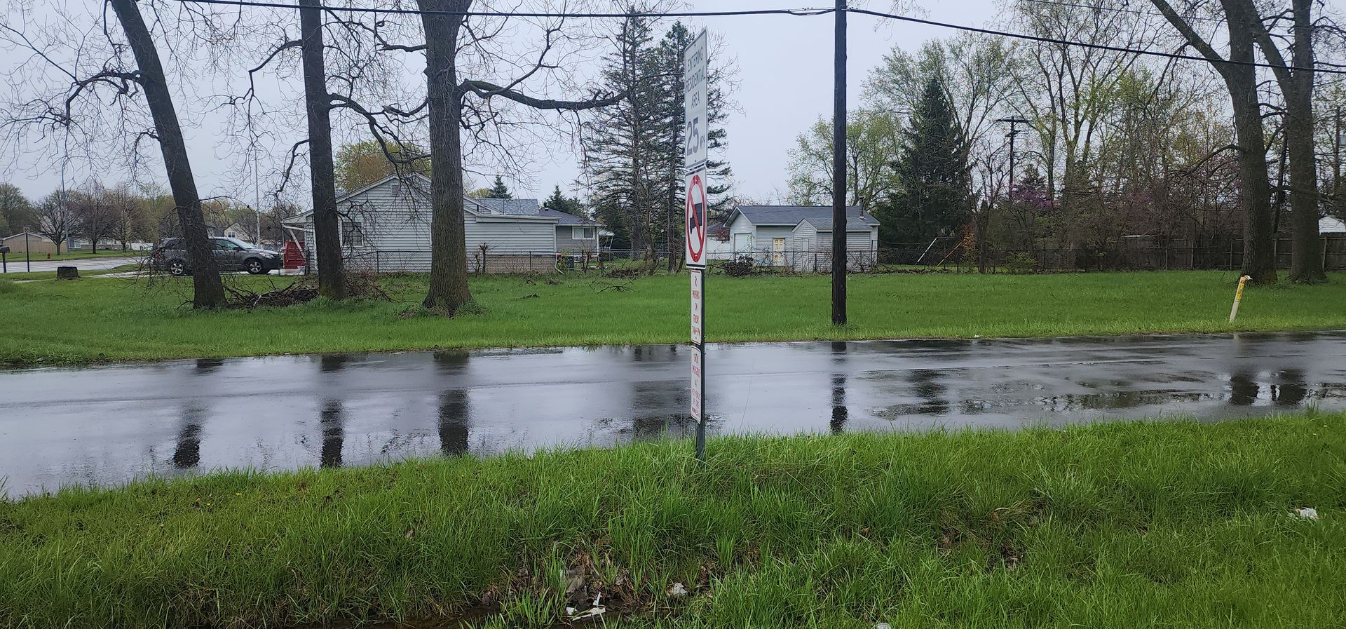 A rainy day scene with a flooded road reflecting trees and houses. Green grass and bare trees in the foreground.