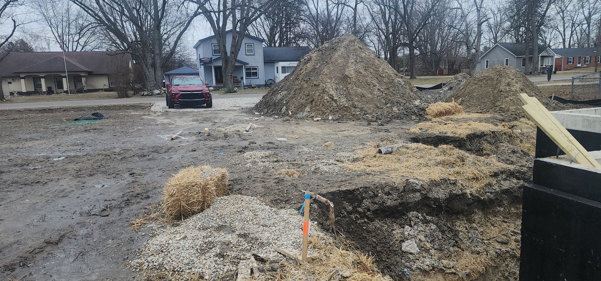 Muddy construction site with piles of dirt, a car, and houses in the background.