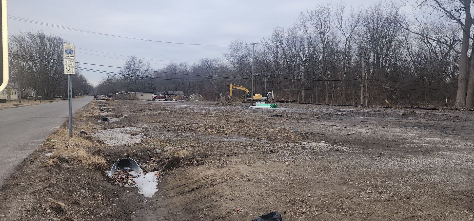 Construction site with excavator and bare trees. Road and sign visible. Gray and brown. Overcast day.