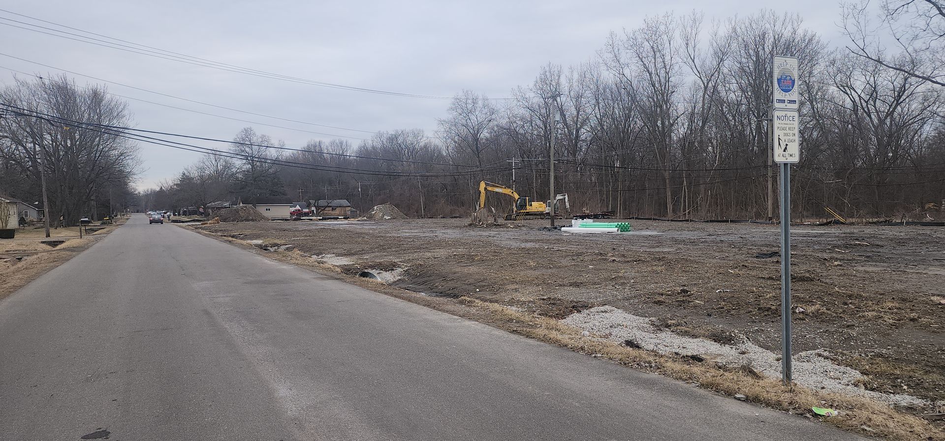 Road with construction site; trees, equipment, dirt, and sign on a cloudy day.