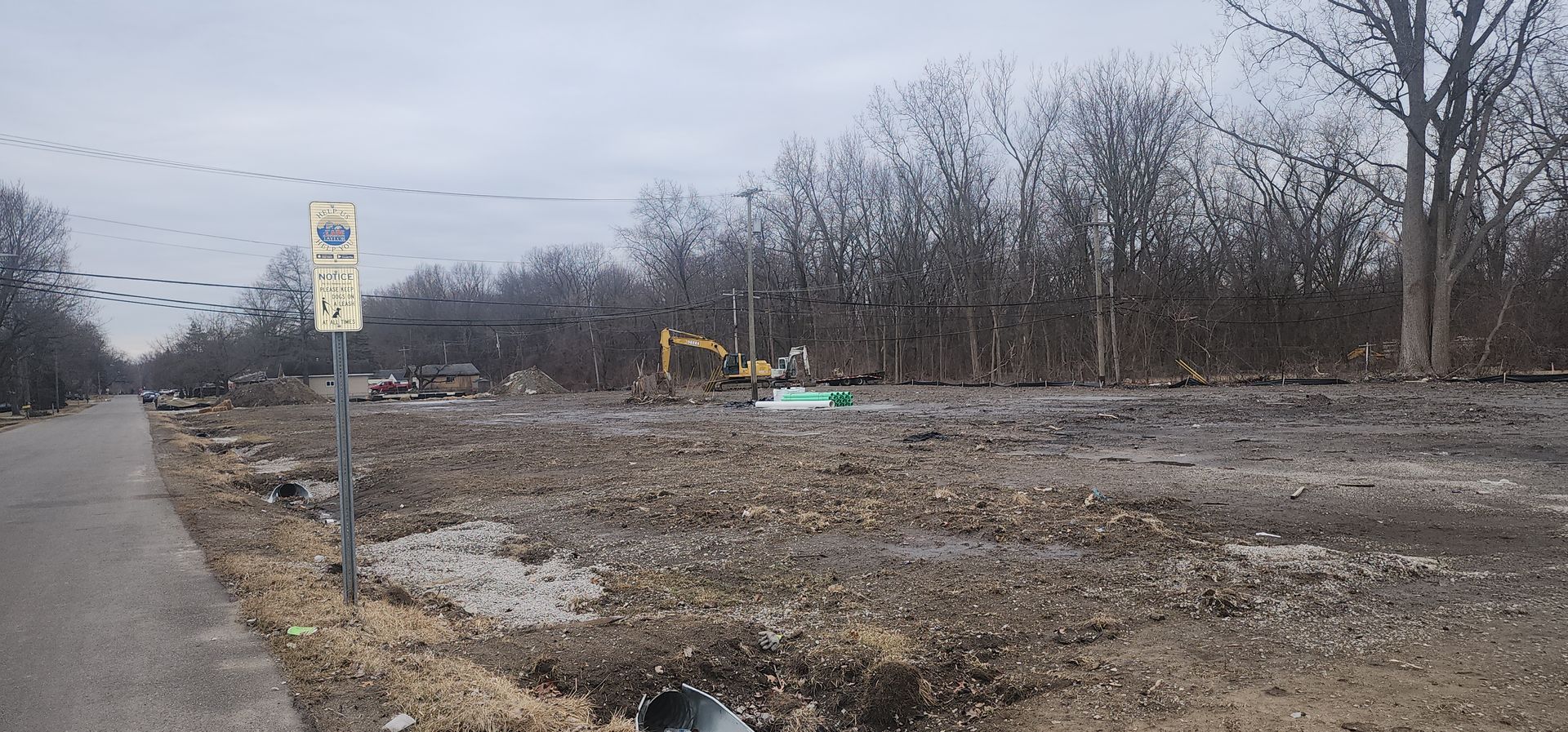 Construction site next to a road with an excavator; bare trees in the background, cloudy sky.
