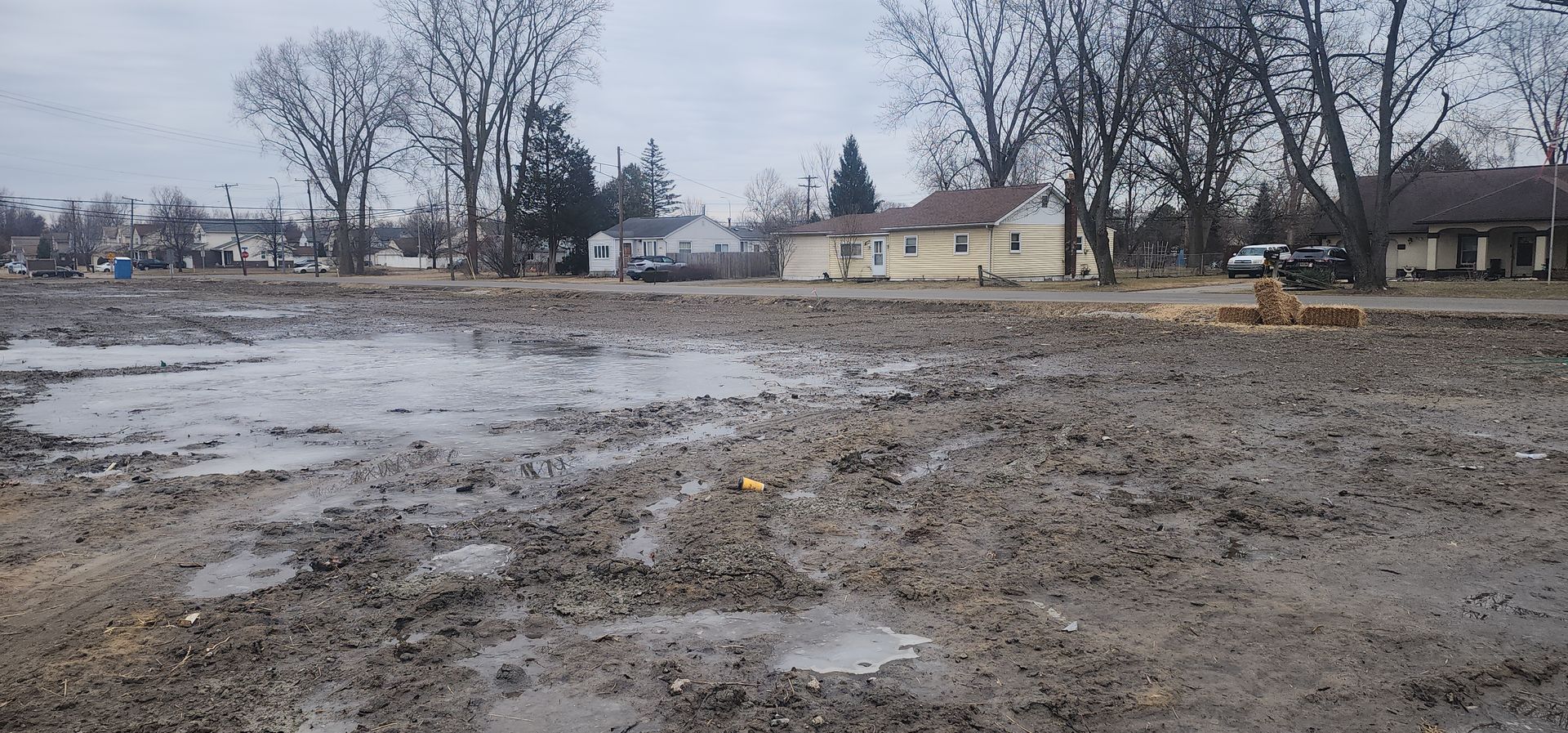 Muddy, bare dirt lot in a residential area with houses and bare trees in the background.