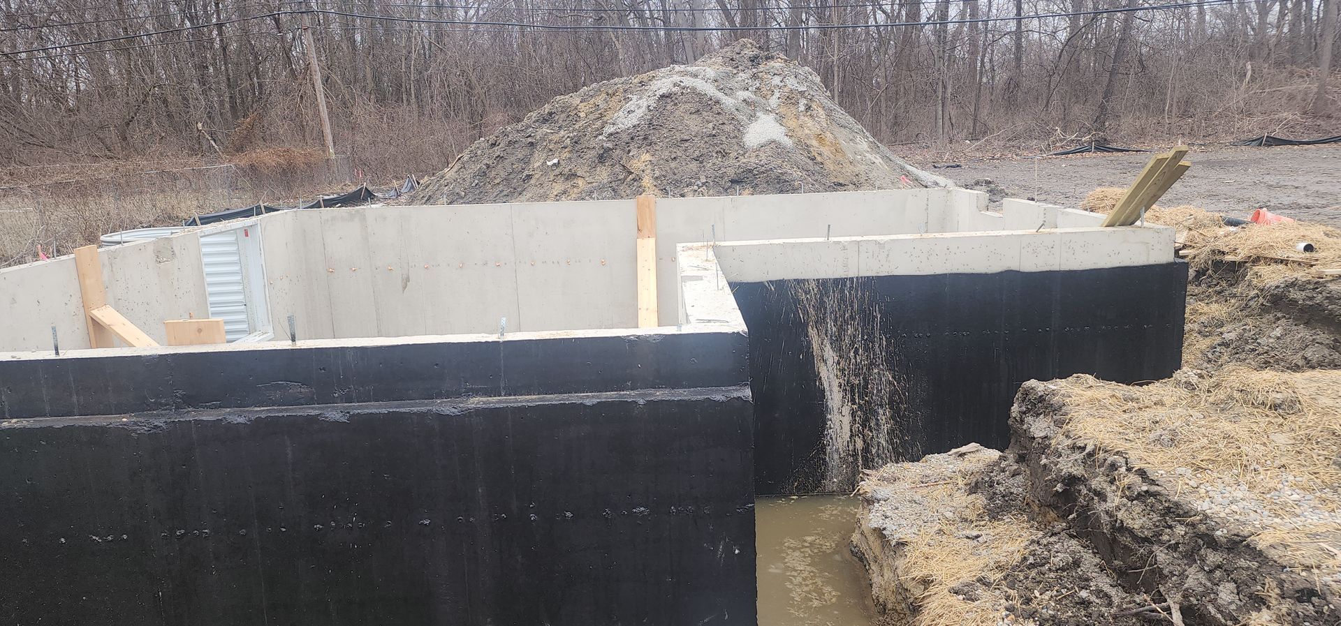 A concrete foundation with black waterproofing is being constructed on a construction site with dirt and trees.