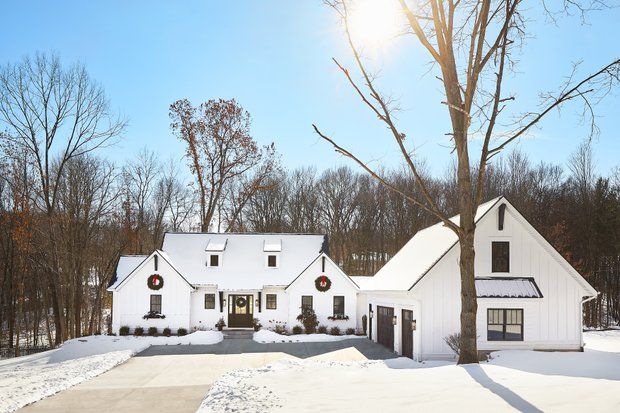 White house with snow-covered roof and driveway, with bare trees in the background. Winter scene, wreaths on doors.