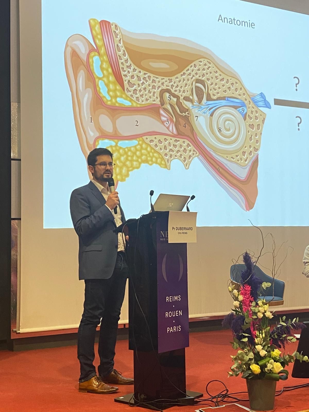 Lors d'une conférence, un orateur, installé à une tribune, présente sur un grand écran un schéma de l'anatomie de l'oreille humaine.