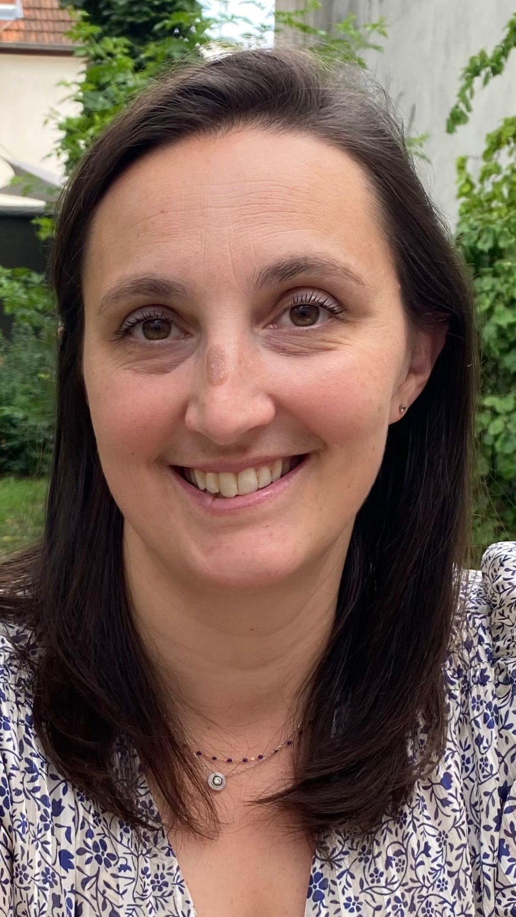 Une personne souriante aux cheveux foncés, vêtue d'un haut à motifs et d'un collier délicat, pose en extérieur devant un décor verdoyant.