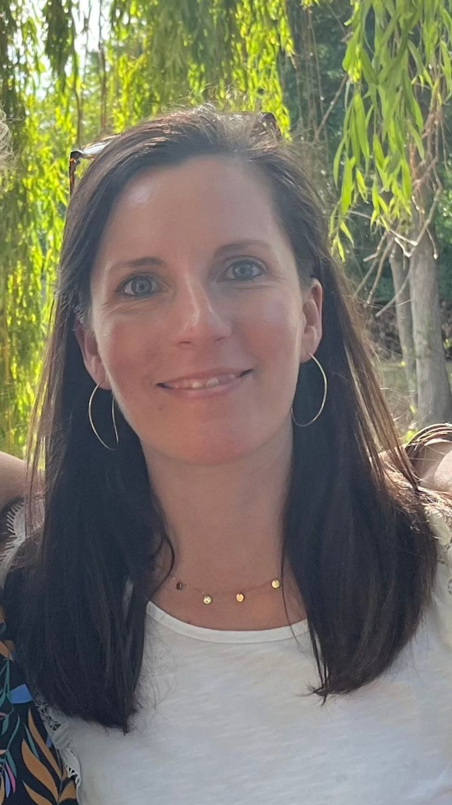 Une personne aux cheveux bruns mi-longs sourit à l'objectif, vêtue d'une chemise blanche et de boucles d'oreilles en or, dans un jardin.