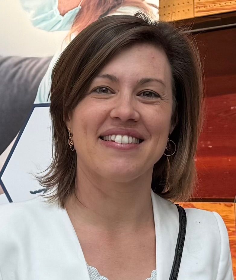 Une personne aux cheveux bruns mi-longs, vêtue d'un blazer blanc, sourit à l'objectif dans un intérieur.