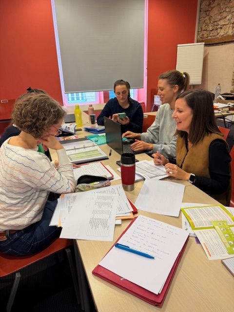 Quatre personnes sont assises autour d'une table dans une pièce bien éclairée, travaillant avec des documents, des ordinateurs portables et une tasse isotherme rouge.