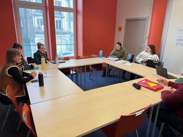Un groupe de personnes est assis autour d'une table en forme de U dans une salle de réunion aux murs orange.