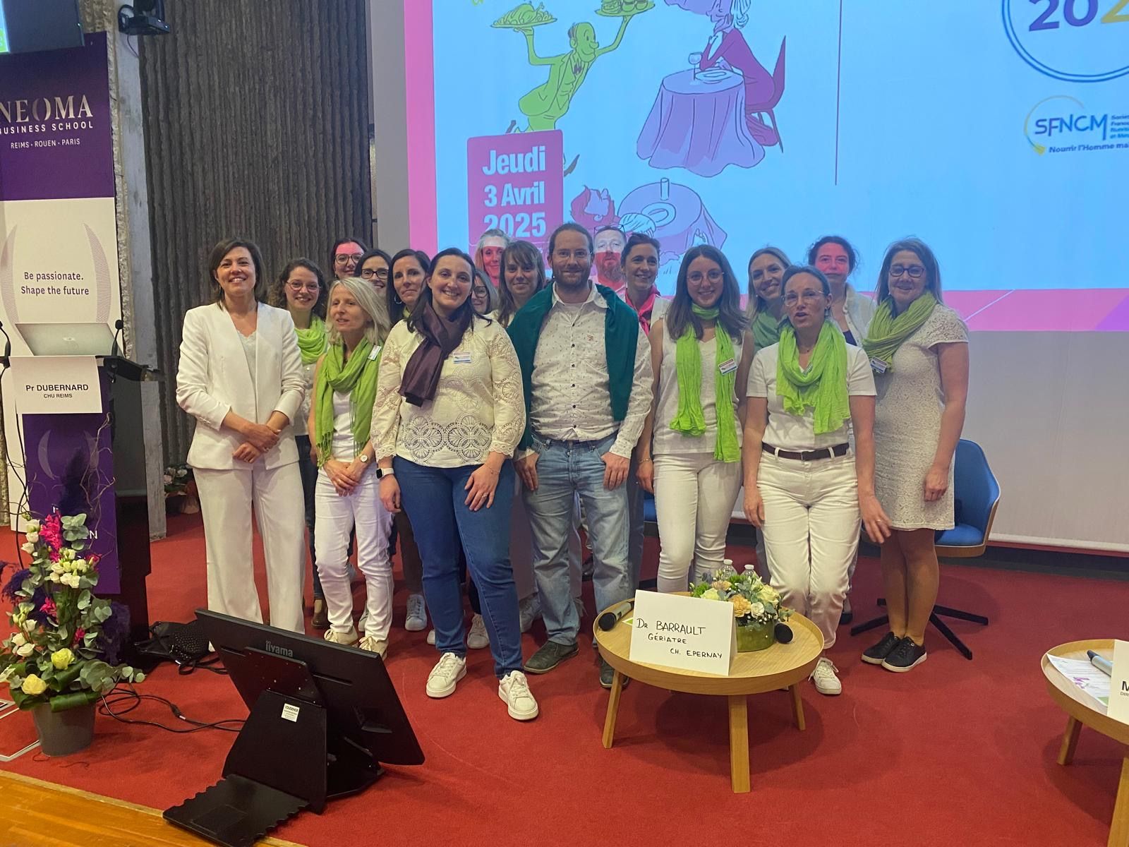 Un groupe de personnes se tenant sur une scène lors d'un événement, beaucoup portant des écharpes vertes, devant un fond de conférence aux couleurs de la marque.