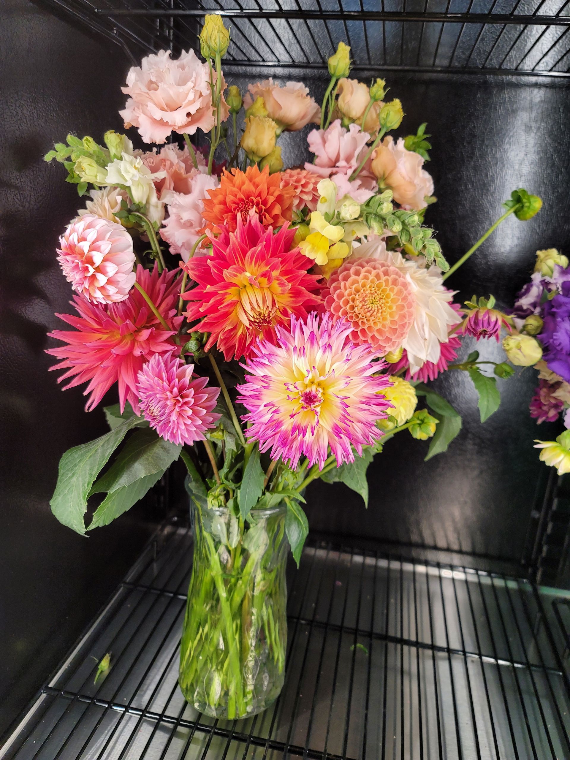 Colorful bouquet of dahlias, lisianthus, and snapdragons in a glass vase on a shelf.