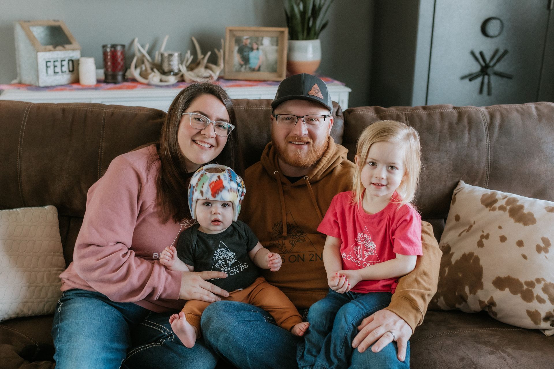 Family of four on a brown couch: parents, baby with helmet, and toddler girl. Smiles, home setting.
