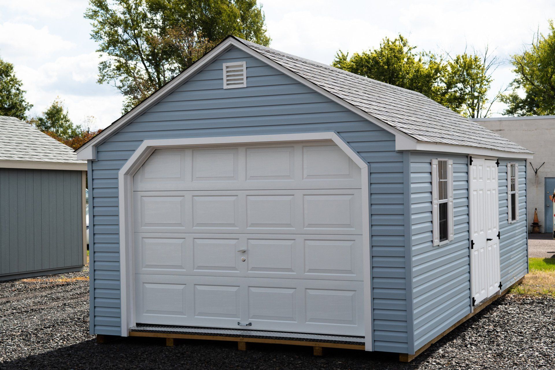 A blue garage with a white garage door is sitting on gravel in front of a house.