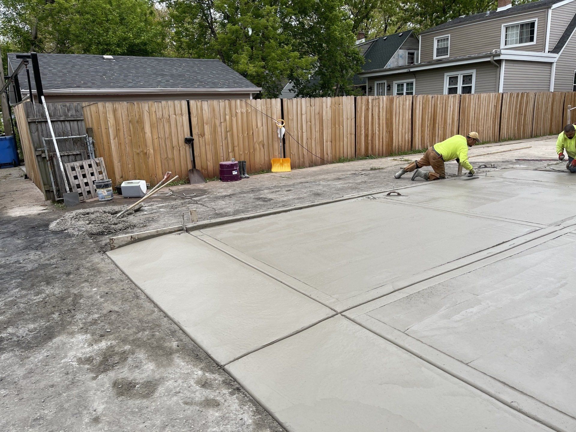 Two men are working on a concrete driveway in front of a wooden fence.