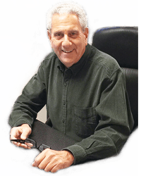 A man in a green shirt is sitting at a desk holding a pair of glasses