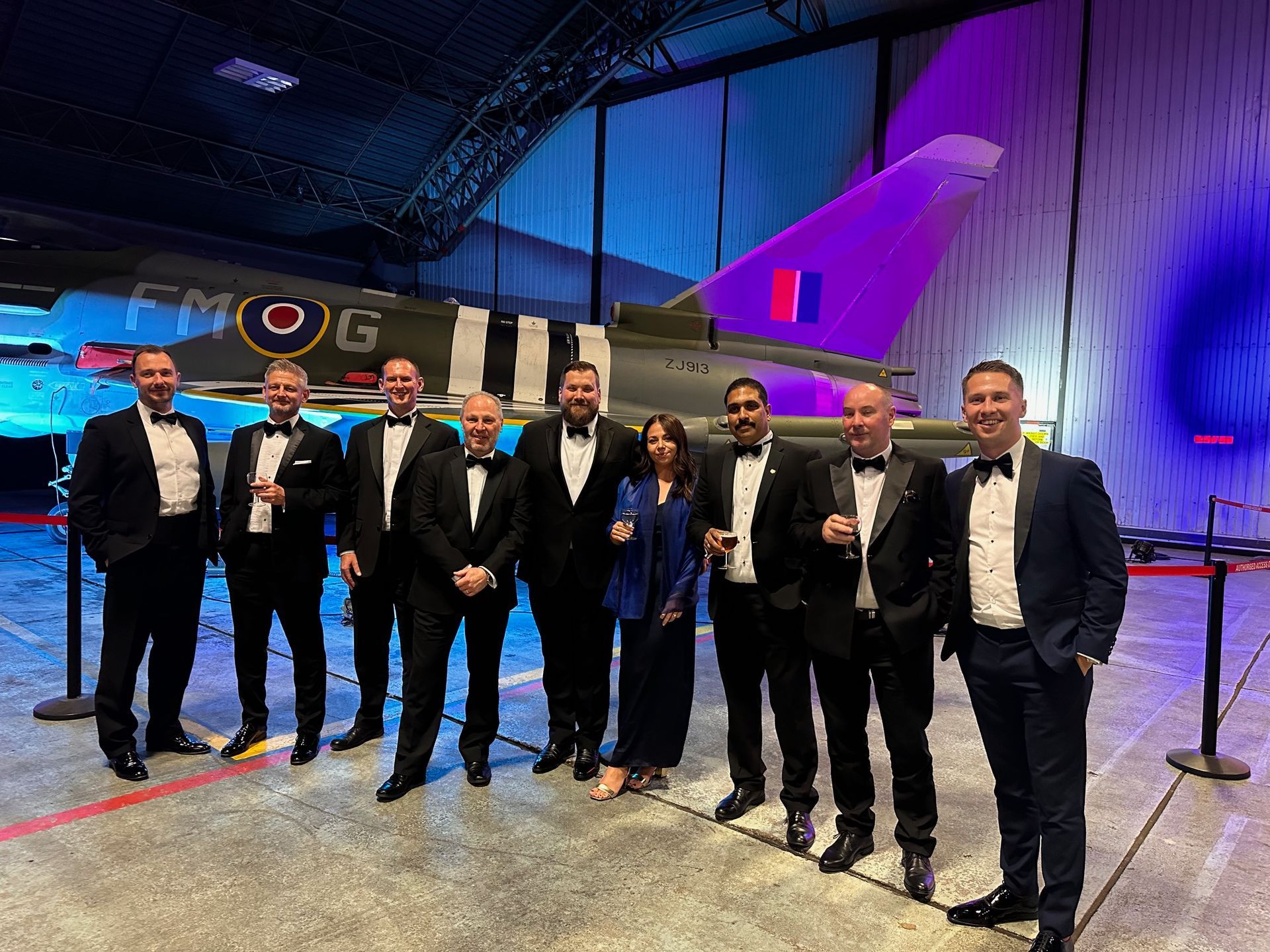 8 men in black tie and one woman in evening dress with an aircraft in the background