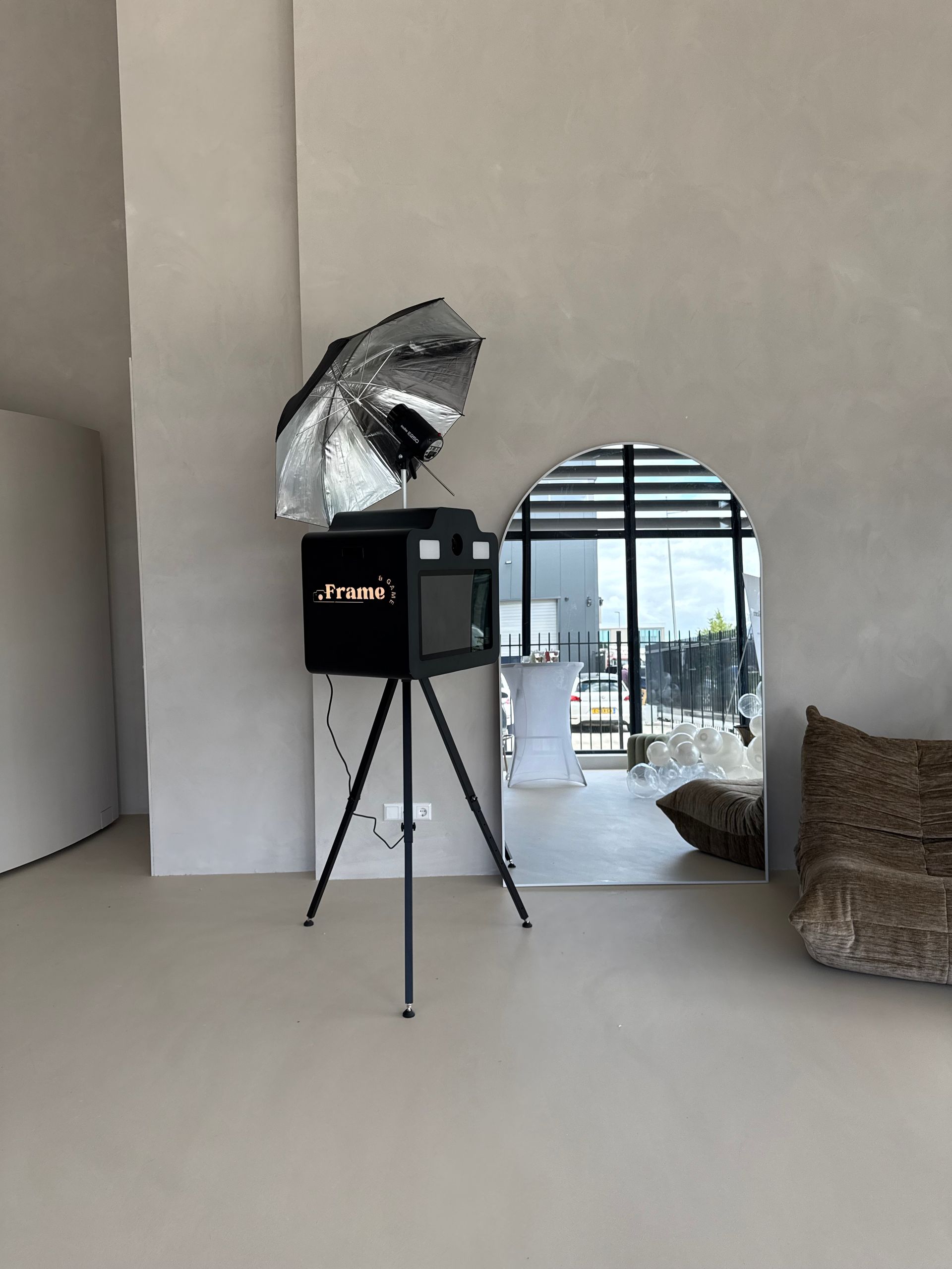 Zwarte fotocabine op statief met zilveren paraplu in een lichte, moderne ruimte bij een groot raam.