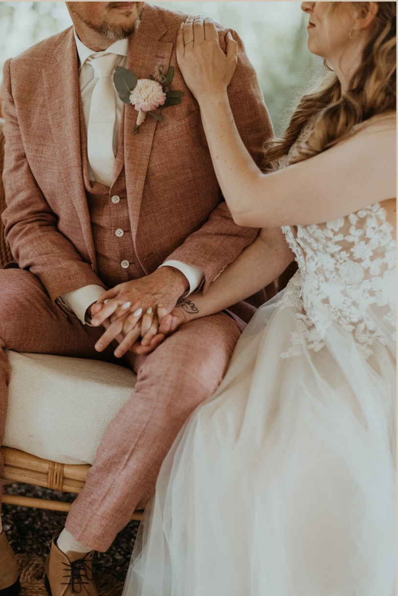 Het bruidspaar zit naast elkaar; de bruid maakt de corsage van de bruidegom recht, beiden in een zachtroze avondkleding.