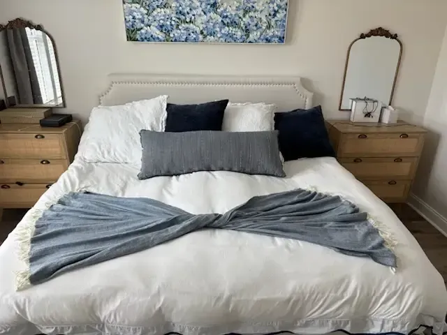Neatly made bed with white linens, navy pillows, and gray throw in a bright bedroom.