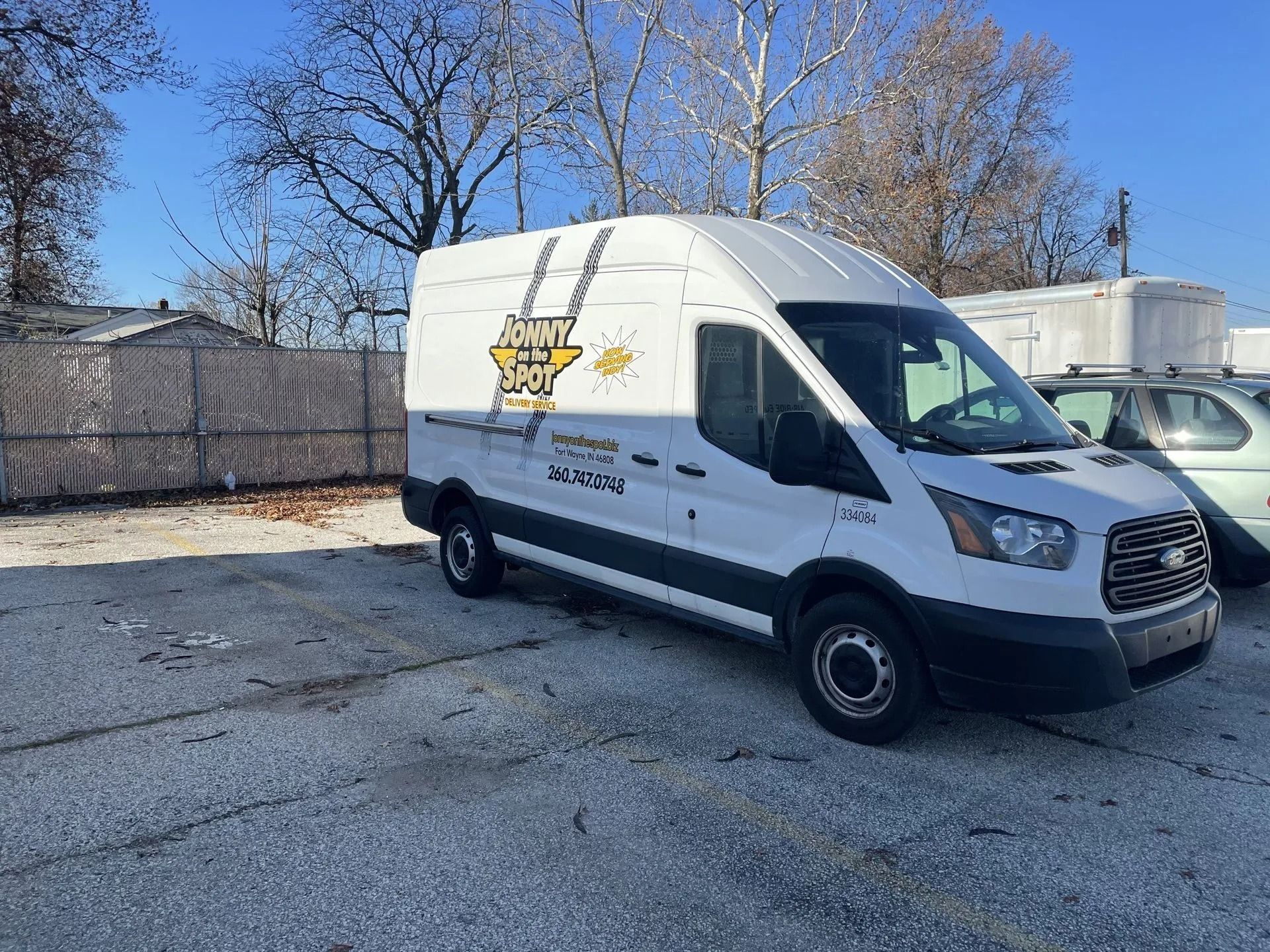 White service van parked on a cracked lot beside a chain-link fence and trees under a clear sky