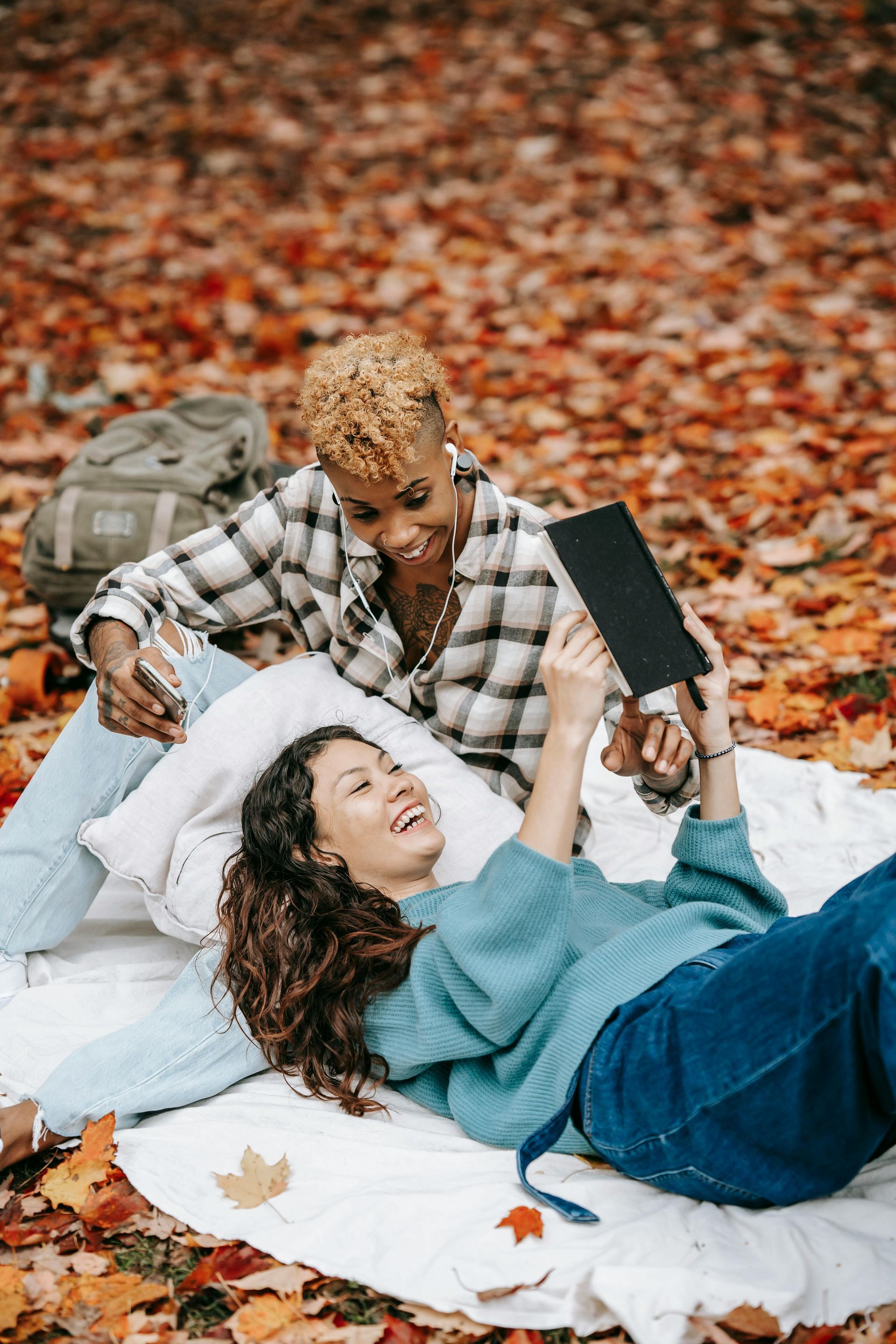 Two people relaxing on a blanket in autumn leaves, one looking at a tablet, the other smiling.