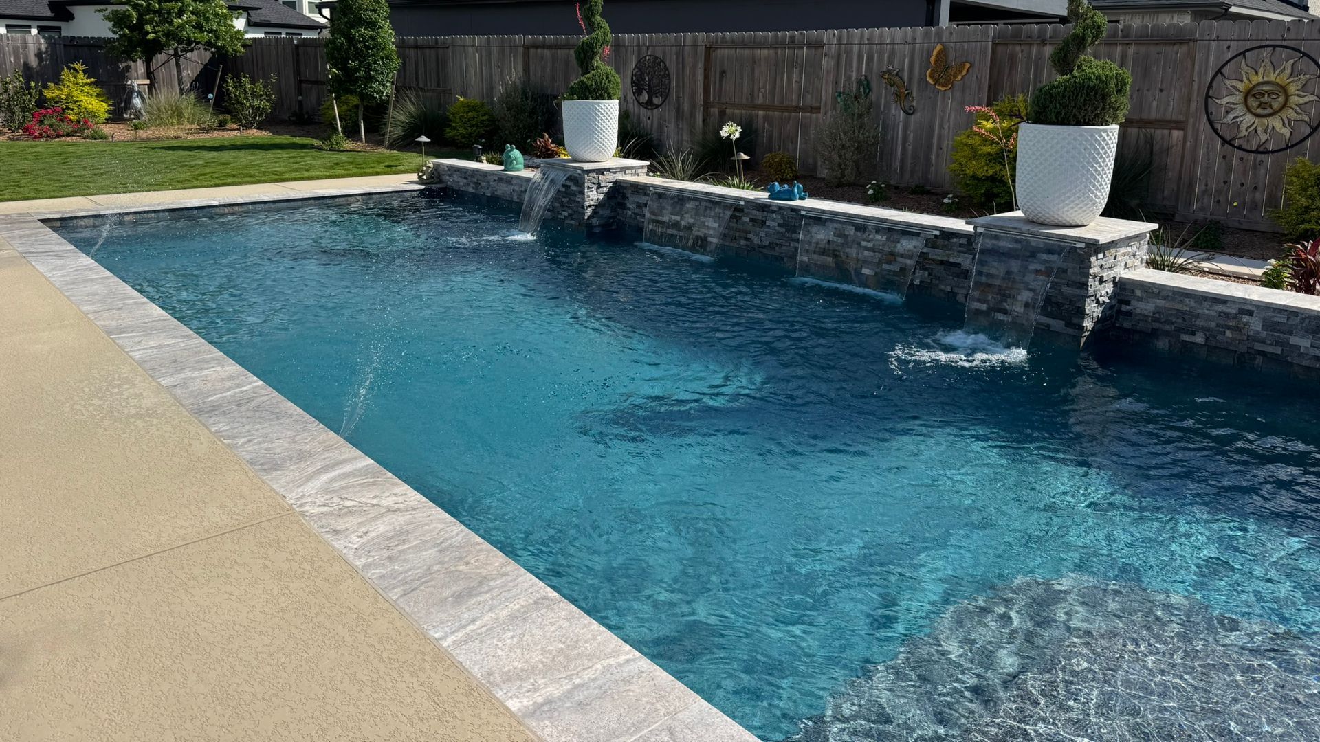 Backyard swimming pool with blue water, stone edge, and white planters along a fence