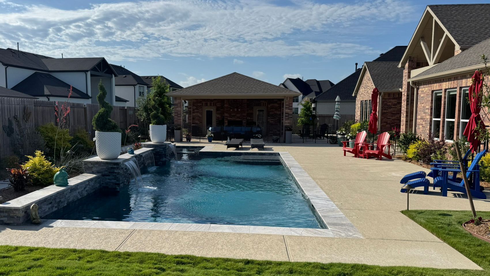 Backyard pool patio with lounge chairs, stone edging, and houses under a clear sky